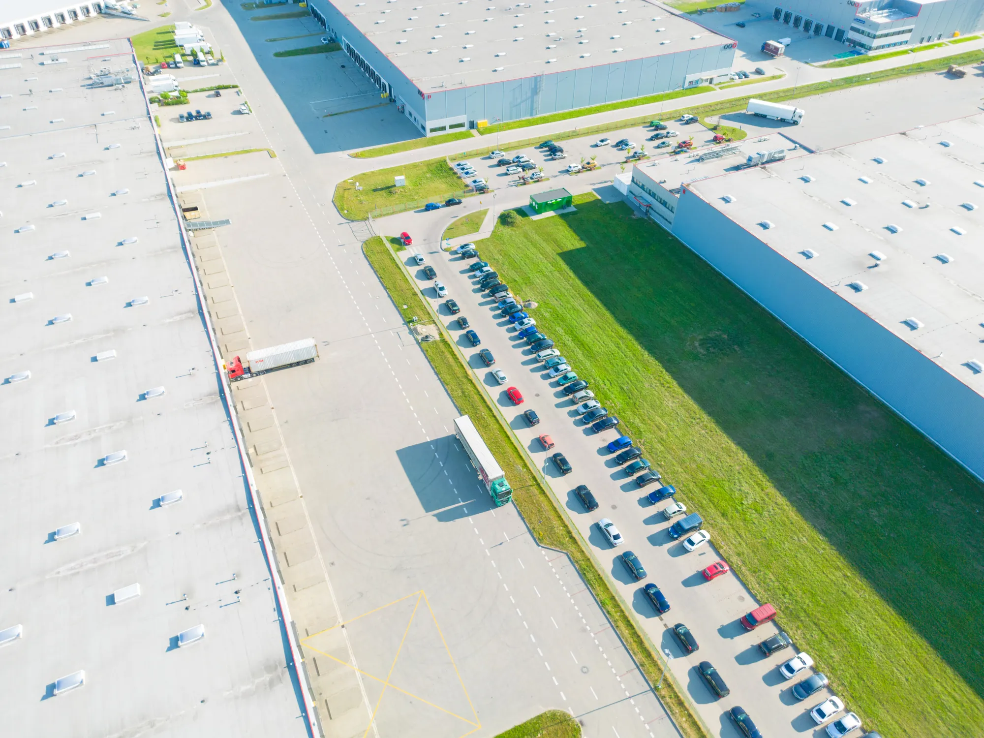 High aerial downward view of a major industrial logistics park complex showing large distribution warehouses, active semi-trucks, and a long row of parked employee cars in Bend, OR