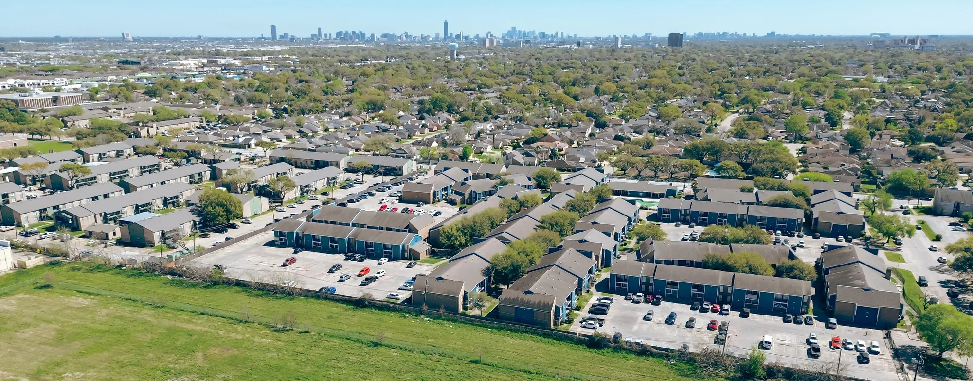 Wide high-angle aerial drone view of a vast suburban garden-style multi-family apartment building complex with parking courts, green lawns, and a distant city skyline corridor in Plano, TX