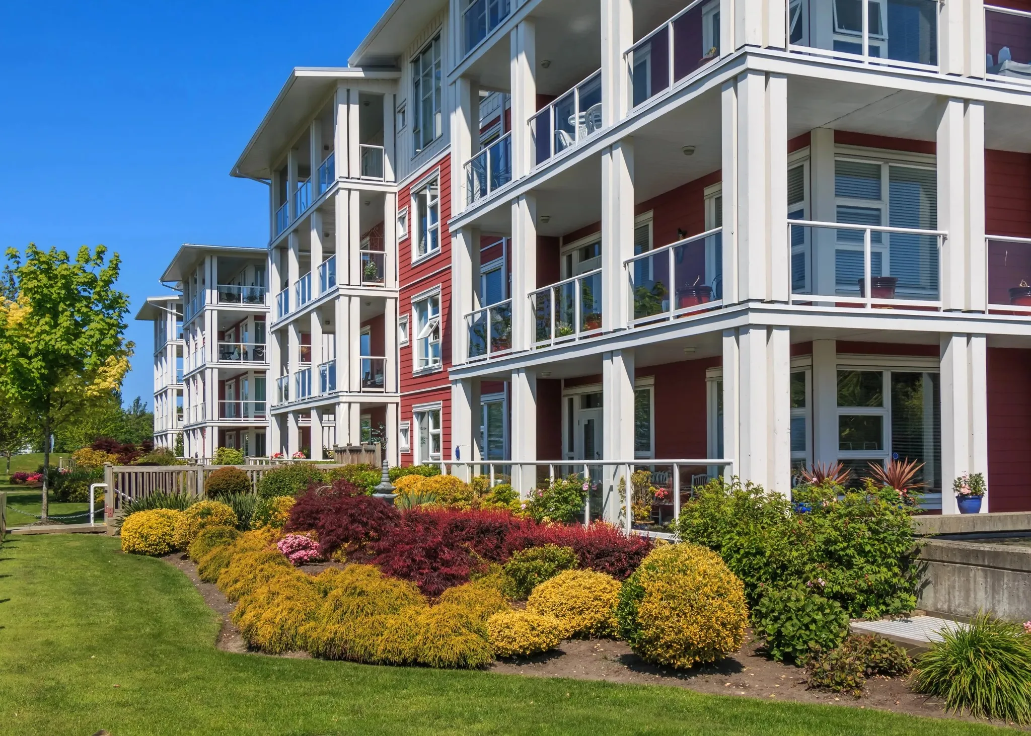 Picturesque wide view of a low-rise coastal style multi-family apartment building with dark red siding, white support pillars, and fully manicured ornamental flower gardens in Halifax, NS