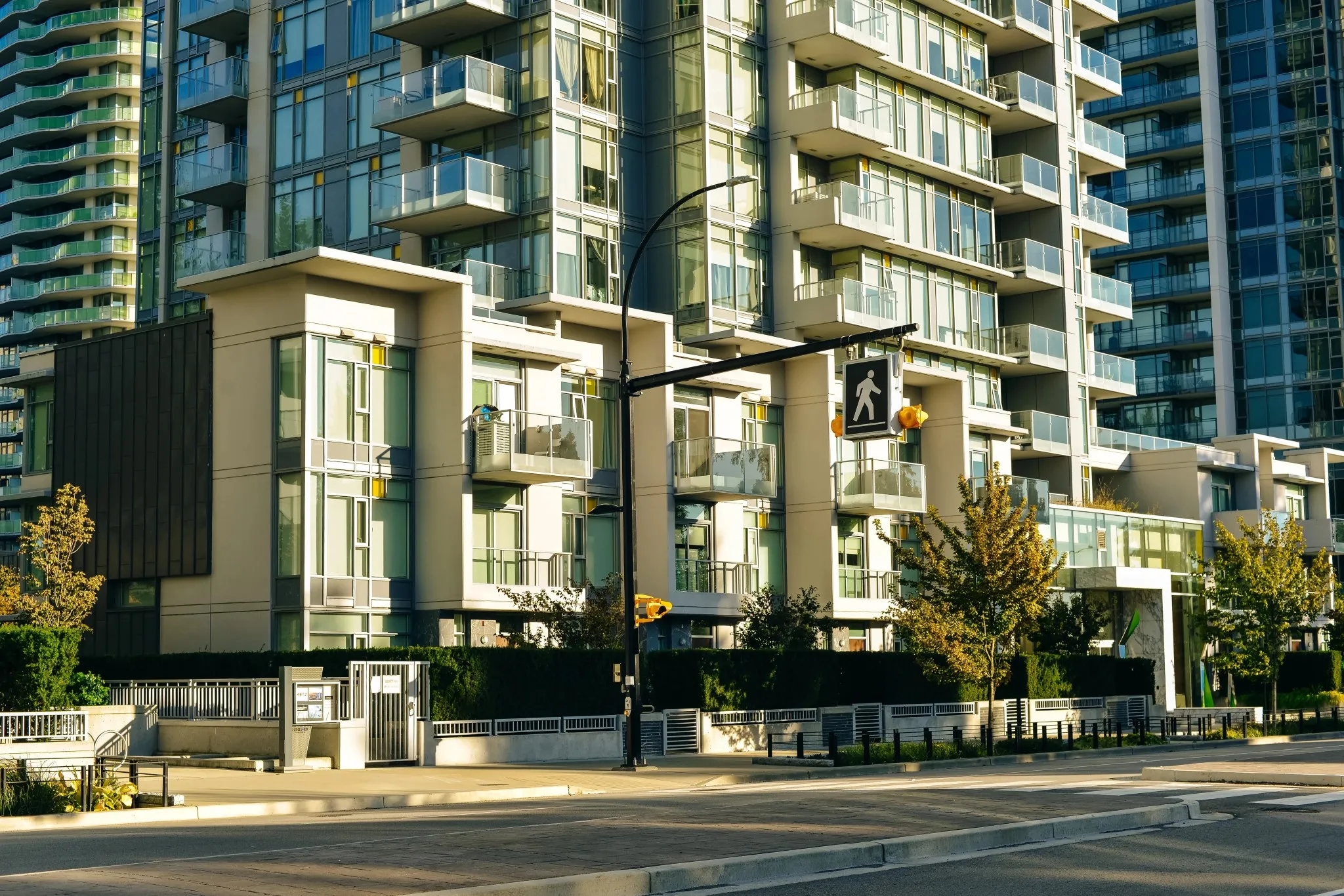 Street-level view of a luxury modern high-rise condominium complex with glass structural railings, a clean concrete crosswalk, and landscaped young trees along the pedestrian path in Toronto, ON