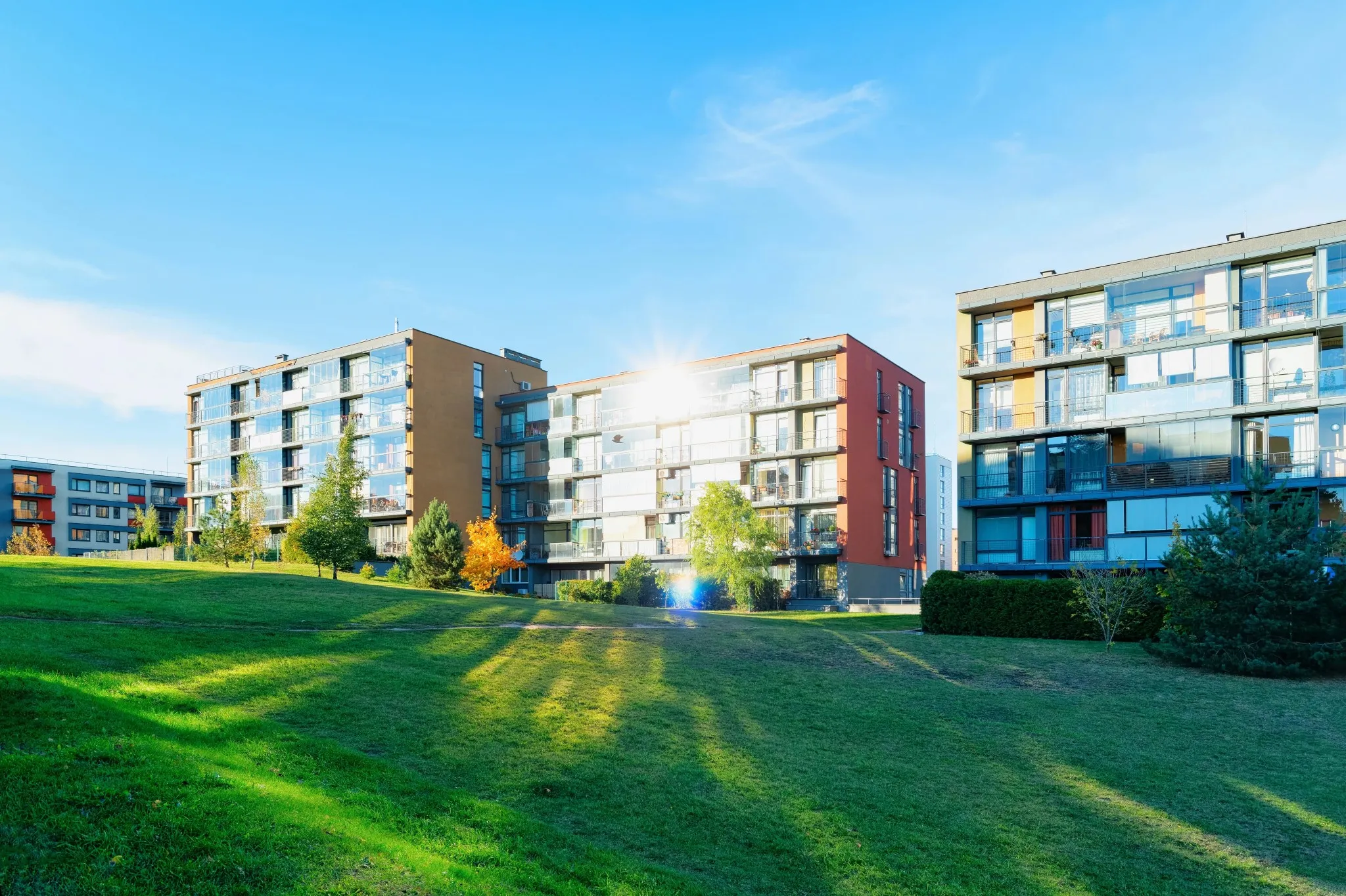 Wide exterior view of a cluster of modern multi-story mid-rise apartment buildings set along a rolling green grass lawn with a bright sunburst flare in the center in Halifax, NS