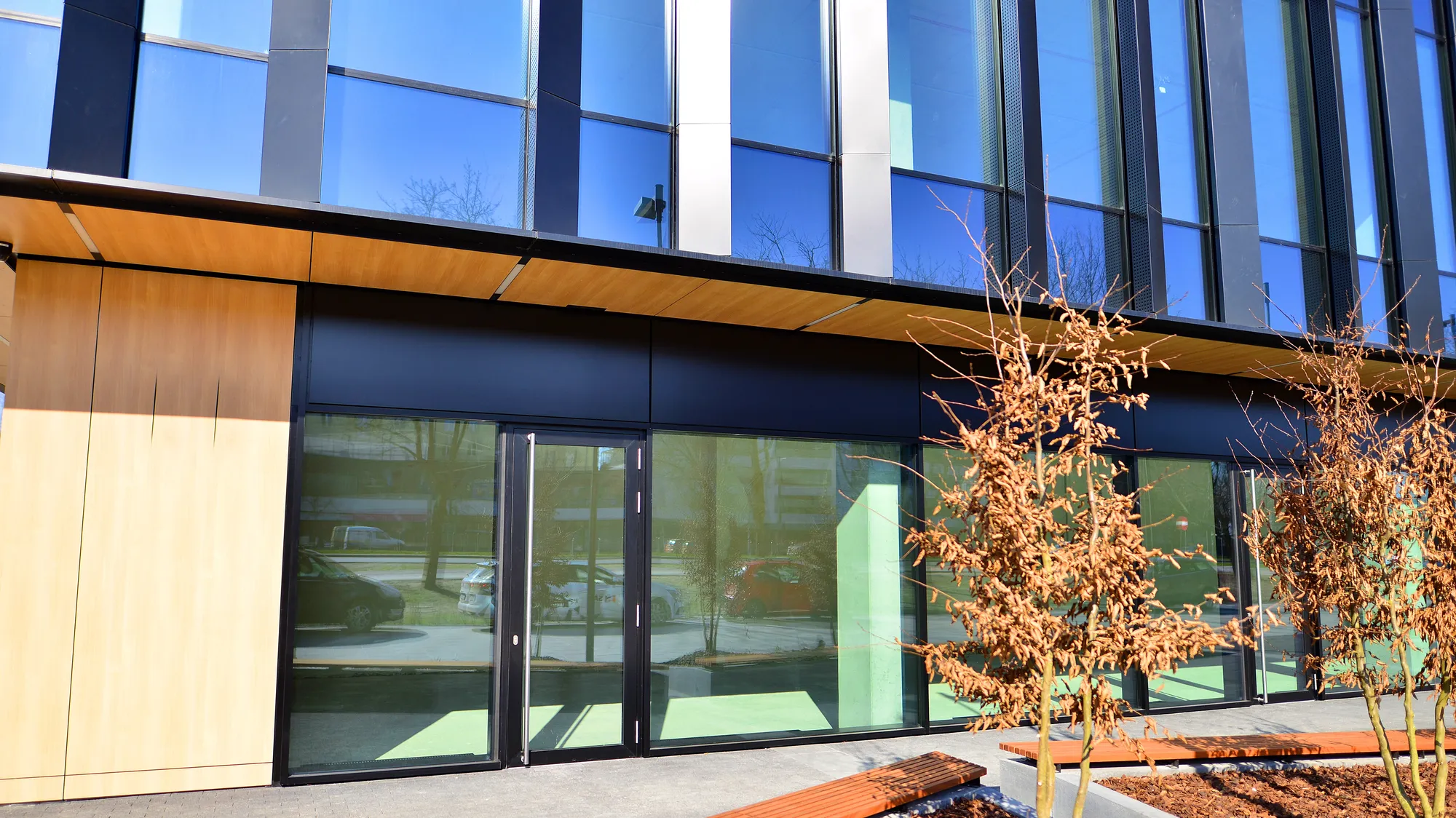 Close-up exterior view of a modern multi-story commercial corporate building featuring a blue reflective glass curtain wall, light-colored wood paneled canopy accents, and landscaped entry benches in Des Plaines, IL