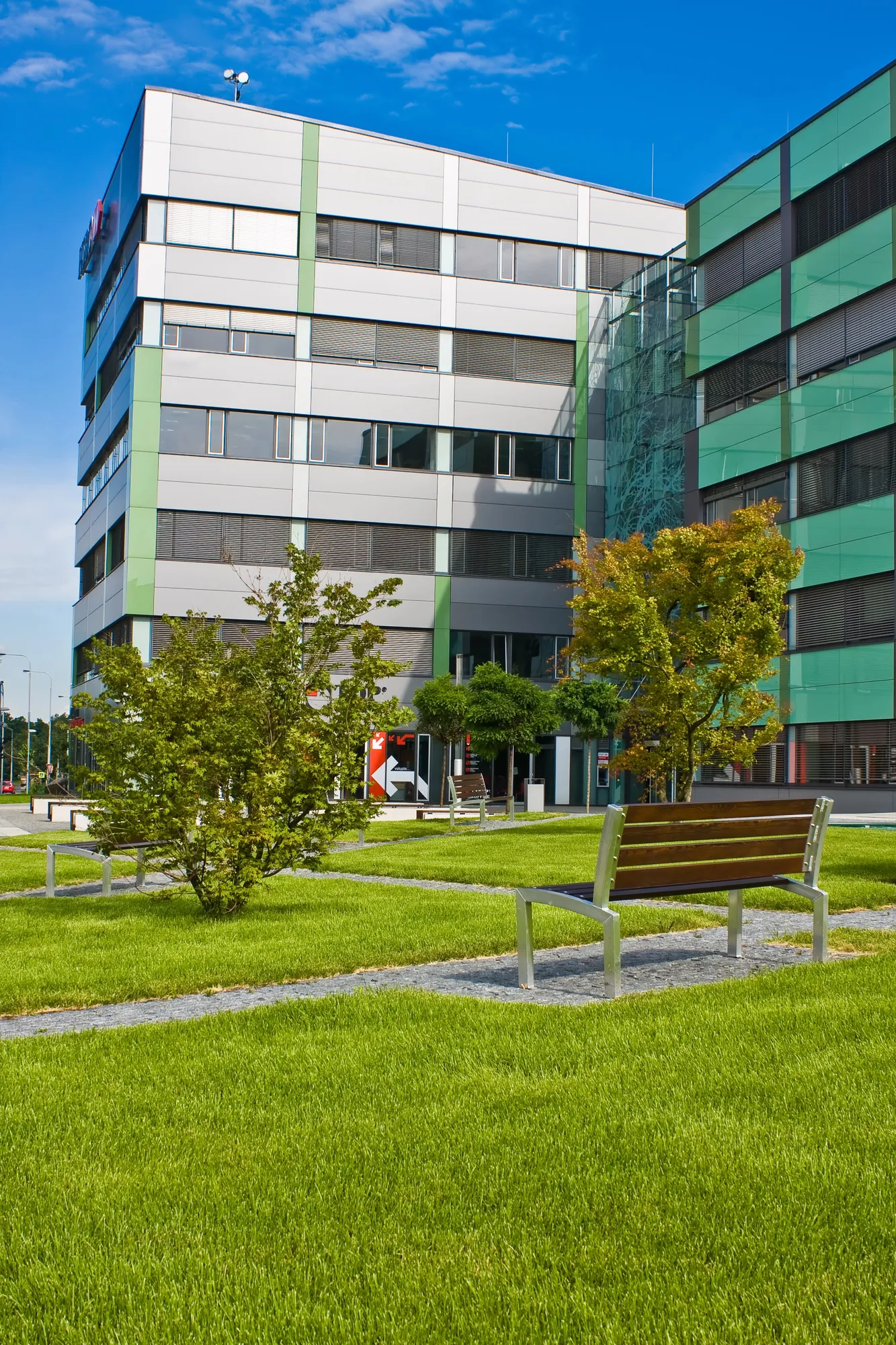 Exterior view of a modern multi-story corporate office building campus with a landscaped green grass courtyard plaza and an outdoor park bench in Aurora, IL