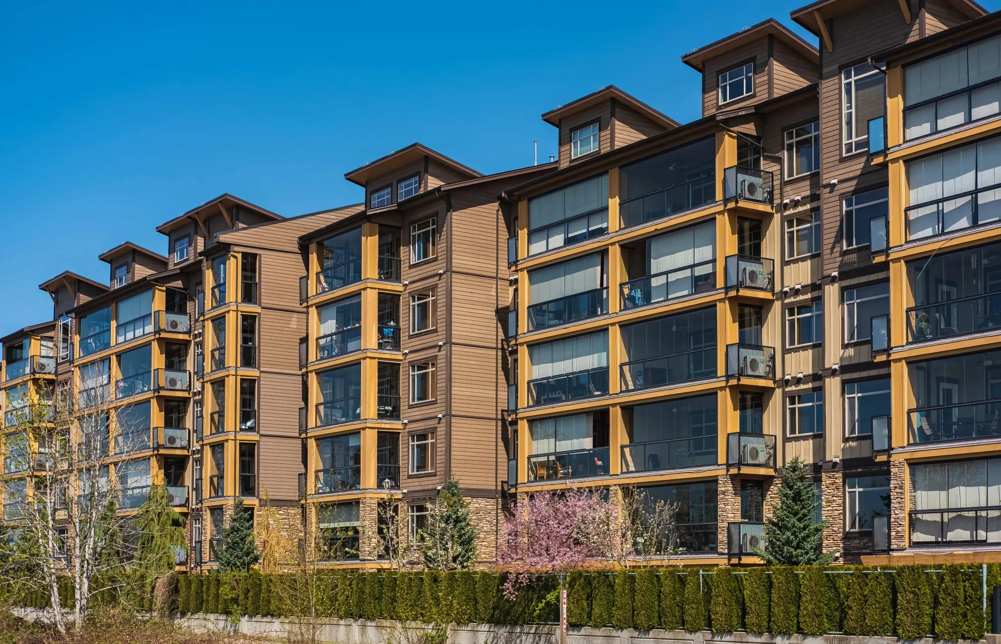 Front exterior architectural view of a modern multi-story timber frame multi-family apartment complex featuring glass balconies and stone accent walls under a clear blue sky in Toronto, ON