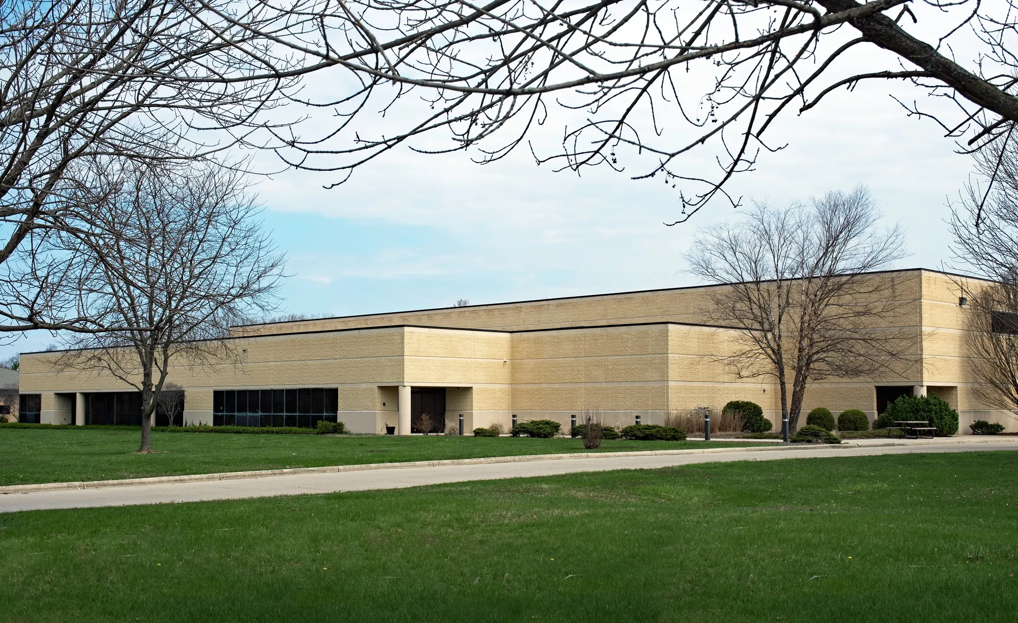 Exterior view of a single-story tan split-face brick commercial office or corporate flex facility surrounded by green grass lawns and trees under a cloudy sky in Chicago, IL